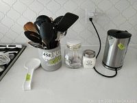 Photo of electric kettle, kitchen utensils in metal container, ceramic spoon rest, and jars on countertop