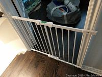White metal safety gate installed in doorway blocking a stairway. Dark wood flooring in background with a bag and shoes visible.