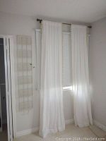 White sheer curtains with fringed edges hanging on a window with a six-pocket white wall organizer nearby.