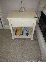 Front view of one white bedside table showing drawer, bottom shelf with books and items, and a lamp on top.