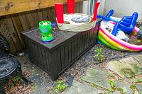 Wide view of the dark brown resin deck storage box outdoors on stone tiles near a fence with children's toys on top and around.