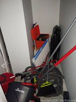 Photo showing a cluster of cleaning items including Vileda mop bucket, broom, black dustpan, multiple grey flexible hoses, and a red metal tool box with tools inside. Black folding chairs stacked against the wall.
