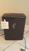 Front view of an Omnitech black paper shredder plugged in resting on a tiled floor with a label 'LOT #11' attached.
