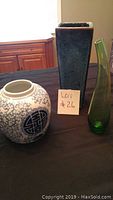 Three assorted vases on a black tablecloth including a blue and white ceramic vase, a tall rectangular blue ceramic vase, and a tall green glass vase.