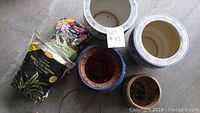 Overview of four plant pots and a bag of potting soil on concrete floor, including large blue and white ceramic pot, smaller brown and beige pots, and soil bag.