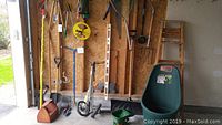 Wide shot showing all garden tools arranged against a wall, including ladder, shovel, wheelbarrow, seed spreader, and other hand tools