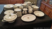 Full view of the entire set displayed on black tablecloth including plates, bowls, cups, saucers, pots, and serving pieces. Pieces have a white base color with a gold rose pattern and gold rim.