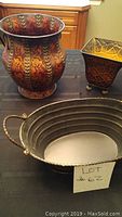 Three decorative metal pots including a large reddish-orange flared pot with leaf pattern, a small square brass pot with textured design, and an oval brass tray with handles photographed on a table.