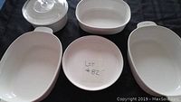 Top-down view of five white CorningWare dishes on black background, including oval roasters and round dish with lid, labeled Lot #82