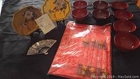Wide layout showing 8 red lacquered bowls with a shiny finish, 6 placemats with chopsticks, and 3 decorative pieces including 2 octagonal plates and a silver fan-shaped item.