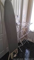Photo showing padded ironing board with grey cover and metal legs standing against a door next to a collapsible clothes drying rack.