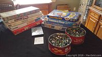 Photo of stacked boxed board games next to two red containers filled with marbles on a black table.