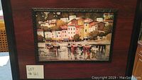 Framed painting depicting boats docked by a waterfront village with buildings and trees.
