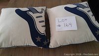 Two square throw cushions laid side by side on wood floor showing guitar designs in navy blue over cream fabric with dark brown sides.