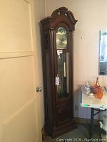 Full view of tall wooden Pearl grandfather clock against wall, showing overall shape and finish.