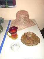 All four items displayed on a white table against a peach wall: pink plastic hat, amberina crackle glass pitcher, carnival glass bowl, and clear glass dish.