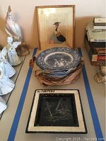 Top-down view of stacked Liberty Blue plates in original brown paper packaging with framed vintage felt artwork and black plaque beneath them.