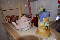 Photo showing ceramic pitchers with deer and nature scene alongside pink floral teapot and amber-yellow glass vases with carnival glass pieces in the background.