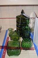 Front and side view of six emerald green glass items on tiled surface, including stacking refrigerator containers, car decanter, candy dishes, and bud vase, with measuring ruler showing approximate size.