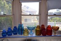 Full view of the entire collection of vintage glassware including blue, amber, and red jars and bowls displayed on a windowsill with bright sunlight.
