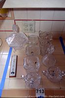 View of multiple covered candy dishes, sugar and creamer, cut glass covered refrigerator dishes, and side pitchers arranged on countertop showing intricate pressed glass details and pointed lid finials.
