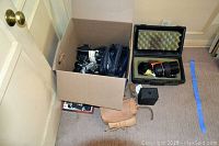 Box on floor containing various camera parts and smaller lenses, a large camera lens in hard case, and a black box nearby.