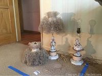 Photo showing set of two vintage ceramic figurine lamps of a boy and girl on white pedestal bases, each topped with ruffled aged beige lace lampshades. Additional separate lace lamp shade placed in front.