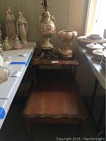Photo showing two vintage Italian lamps with ornate ceramic bases and cherub and floral relief designs, placed on a wooden two-tier side table with scalloped edges.