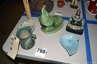 Photo showing multiple vintage ceramic and glass items on a flat surface: a green jug, a hen figurine, a blue dish, a figurine in a dark base, and a reddish vase.
