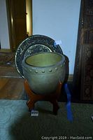 Front view showing the round metal planter with rivet decoration and wooden stand, plus the large embossed metal tray in the background.
