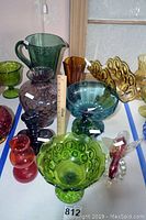 Full assortment of vintage glassware including green Moon and Star nappy bowl, purple Crown Bottle, blue glass compote, small colored vases, and glass bird.