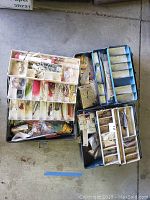 Three plastic tackle boxes with fishing accessories inside, shown in garage or similar environment.