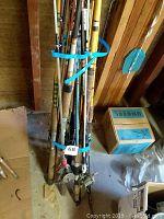 Bundle of multiple fishing rods tied together with blue straps showing variety of rod types and materials, leaning against wooden wall.