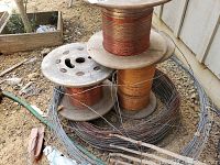Three partial rolls of copper wire on wooden reels stacked outdoors on dirt ground with surrounding metal wire and debris.