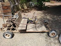 Side view of the vintage homemade go-kart showing metal frame, wooden bench seat, rear engine and front wheels.