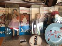 Photo of three Norman Rockwell character dolls in original packaging with protective boxes visible, showing a girl, a boy named Davey, and a male figure.