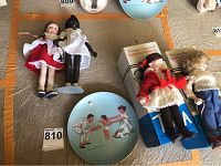 Overview of four porcelain dolls and Norman Rockwell decorative plate shown on a flat surface with scale marker.