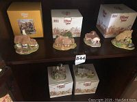 Six miniature Lilliput Lane houses displayed on a wood shelf, each with original boxes behind them. Houses feature various English countryside styles, including cottages and a church building.