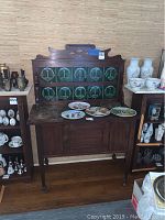 Front view of antique buffet showing decorative green tiled back splash and wooden cabinet below with some decorative plates on top.