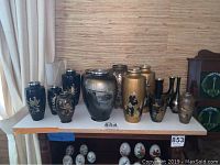 Fifteen vintage Japanese enamel vases grouped on a white shelf showing different sizes, colors, and decorative motifs including floral and landscape scenes.