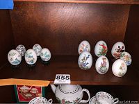 Photo showing twelve hand-painted decorative eggs with detailed artwork, some standing on small black stands, arranged on a wooden shelf.