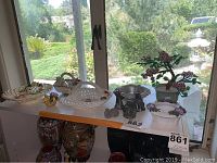 Display of animal figurines, white woven clay baskets, Asian glass bonsai tree, ceramic planter with purple flowers, and metal bowl on shelf in front of window.