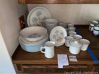 Full set of dishes for eight displayed on wooden surface, arranged with plates, bowls, cups, saucers, creamer, and sugar bowl with lid.