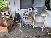 Three wooden crates including 7UP crate, black metal mailbox on white metal stand, six rustic wooden bird houses on white round metal table, silver U.S. MAIL mailbox on cream wooden table, and metal stool partly visible.