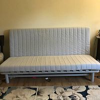 Photo showing white sofa cum bed with a padded cover and iron frame, placed in room with wooden floor and patterned rug.
