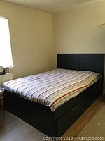 Full view of the black queen bed frame with paneled headboard in a room. Striped mattress cover is on the frame, but mattress is optional and not for sale. Storage drawers visible under bed are broken.