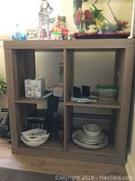 Photo showing a wooden square 4-cube storage organizer with various items on it and inside compartments, against a wall with framed artwork.
