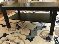 Photo showing black rectangular coffee table with lower shelf and four legs on a floral patterned carpet.