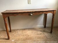 Photo of a standalone wooden table with a laminated top. The table has a medium brown finish, four turned legs, and a single drawer with a brass-style handle. The top surface and legs show some signs of wear and use.