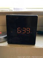 Black digital table clock showing the time 6:39 PM in orange LED digits, set against a window ledge background.
